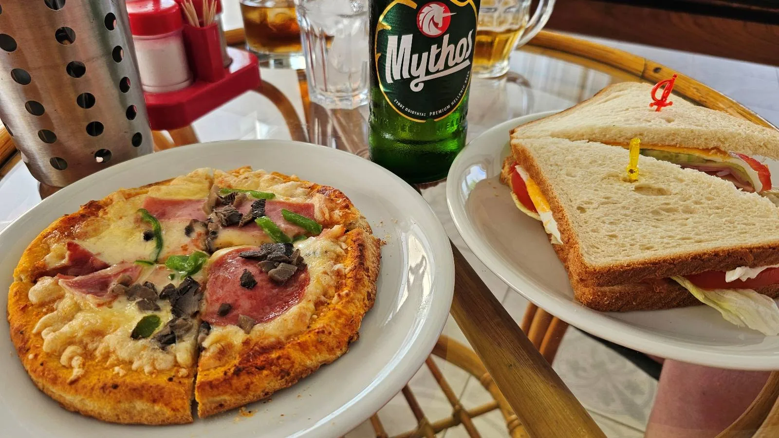 A small pizza with ham, mushrooms, and herbs on a white plate sits next to a sandwich with lettuce, tomato, and cheese, along with a green bottle of Mythos beer on a table.