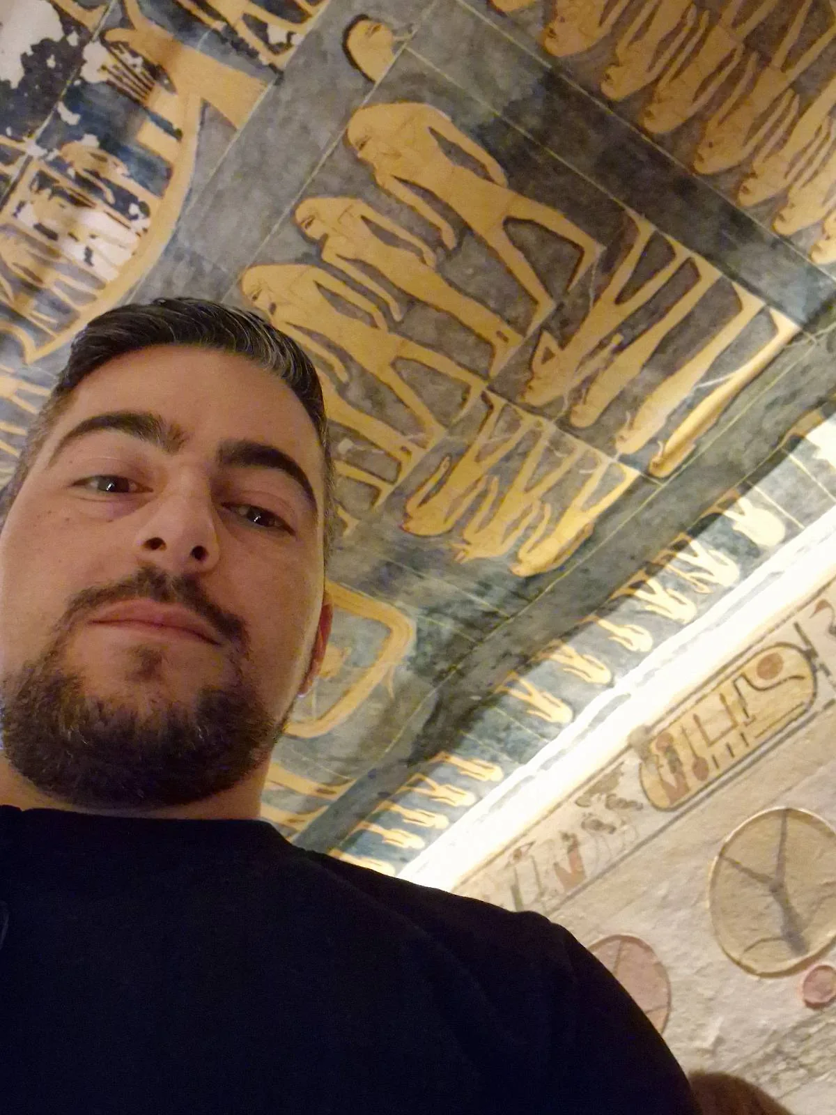 A person with a beard takes a selfie inside an ancient structure, featuring detailed wall and ceiling art with Egyptian-style figures in gold and blue tones. The person gazes at the camera, and the art is prominently displayed above.