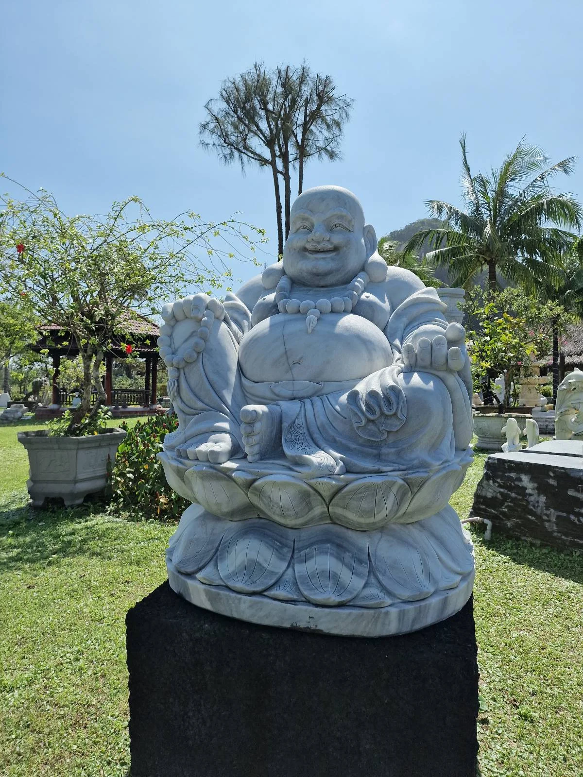 A marble statue of a laughing Buddha sits on a pedestal in a garden under a clear blue sky, surrounded by greenery and trees.