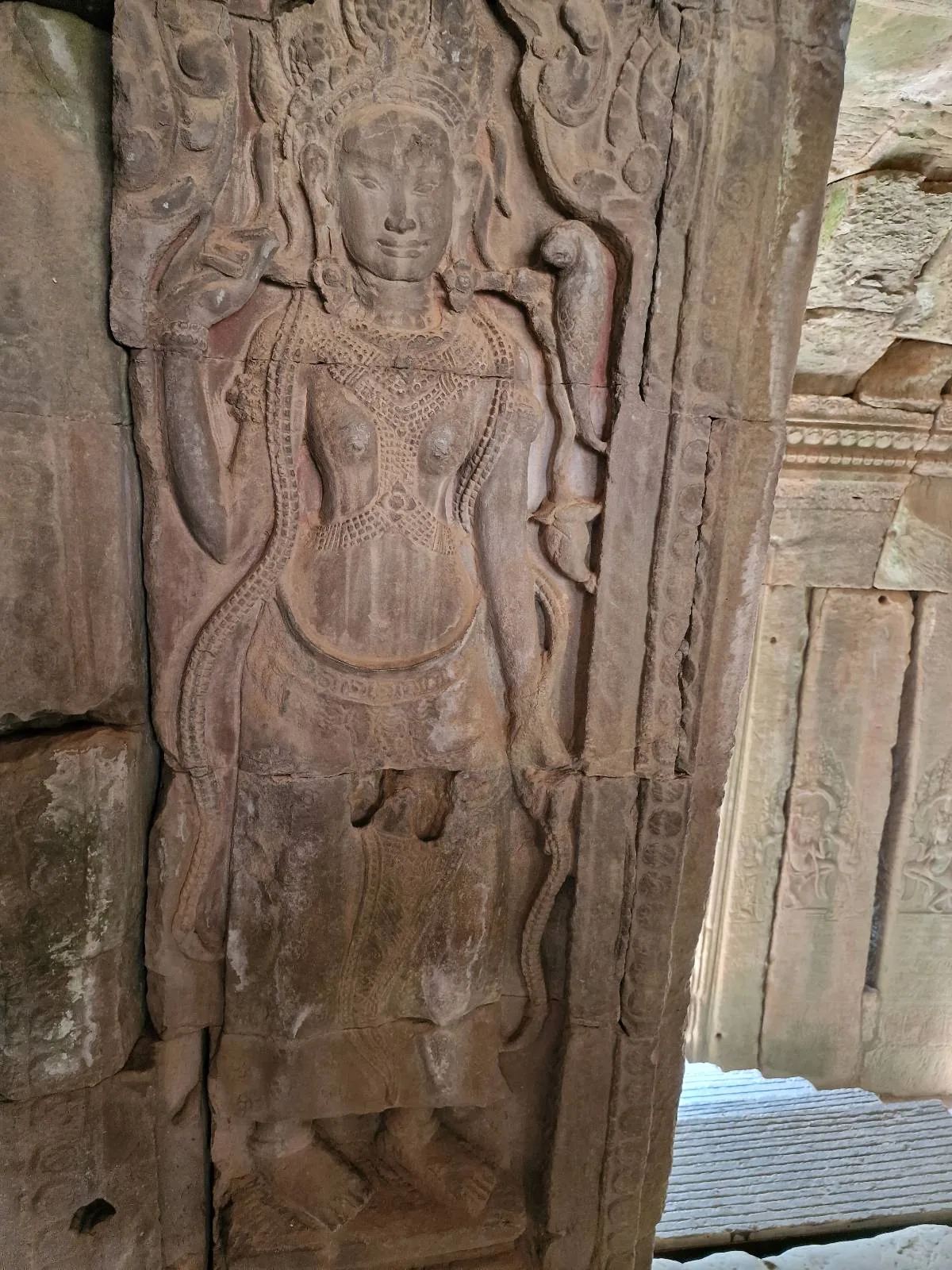 A stone carving of a standing female figure, adorned with jewelry and a headdress, is etched into an ancient temple wall with intricate details.