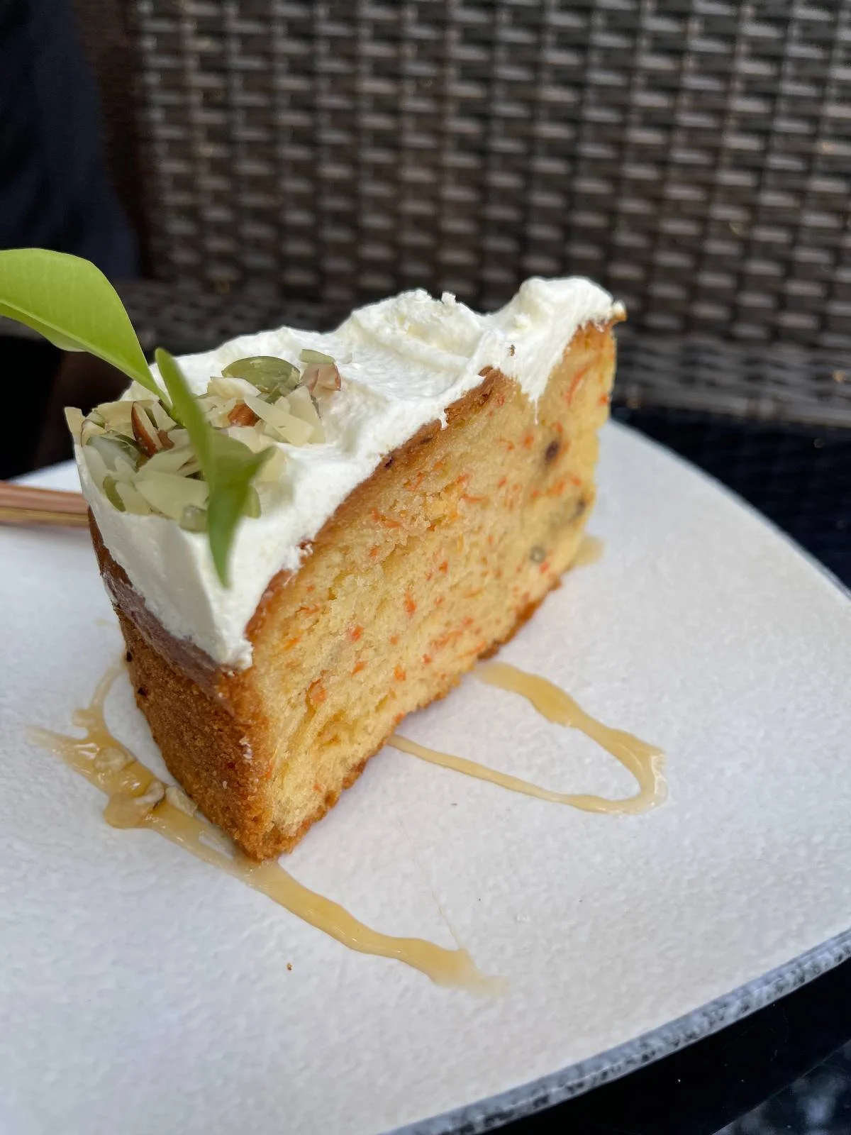 A slice of carrot cake with white frosting, garnished with a small green leaf and a sprinkle of nuts, sits on a white plate. A drizzle of honey decorates the plate beneath the cake. The background features a woven texture.