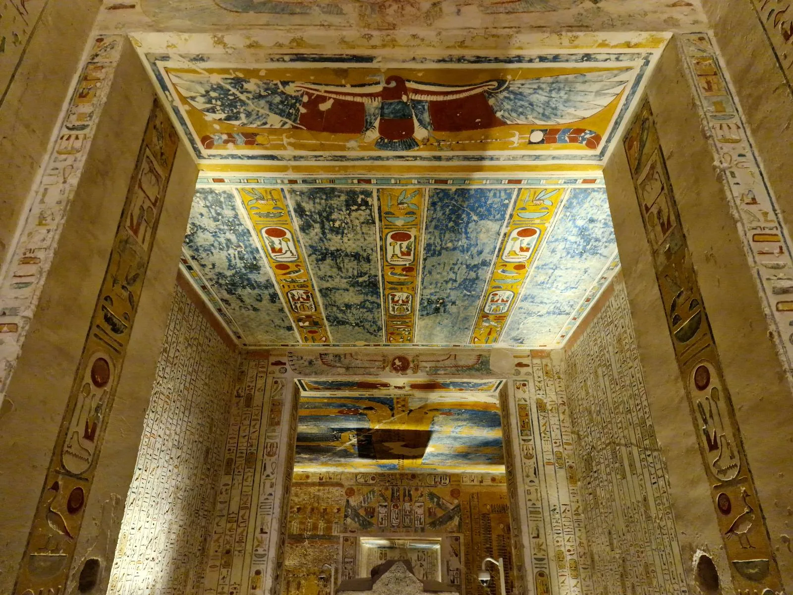 A richly decorated room with ancient Egyptian hieroglyphs and paintings covering the walls and ceiling, including a large blue and red depiction of a figure with outstretched wings on the ceiling.