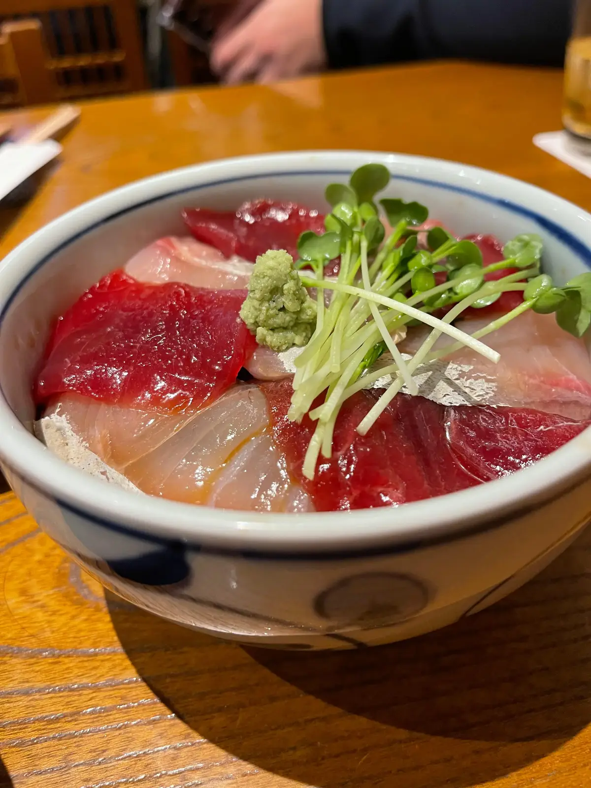 A bowl of sashimi donburi with slices of raw fish, wasabi, and microgreens on top, served on a wooden table.