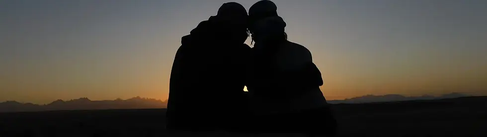 Silhouette of two people sitting closely together during sunset, with a mountain range in the background.