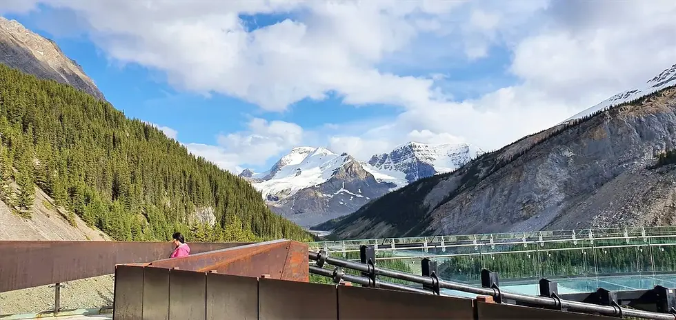 A person in pink walks along a scenic pathway with mountains, forests, and a river under a blue sky.