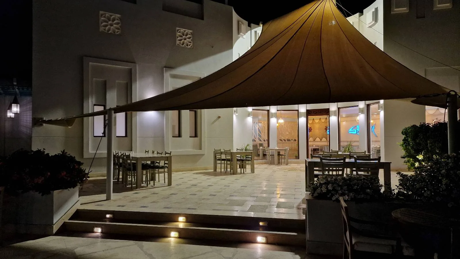 Outdoor patio of a building at night, featuring a large triangular shade sail, lit steps, and dining tables and chairs arranged neatly on the stone floor.