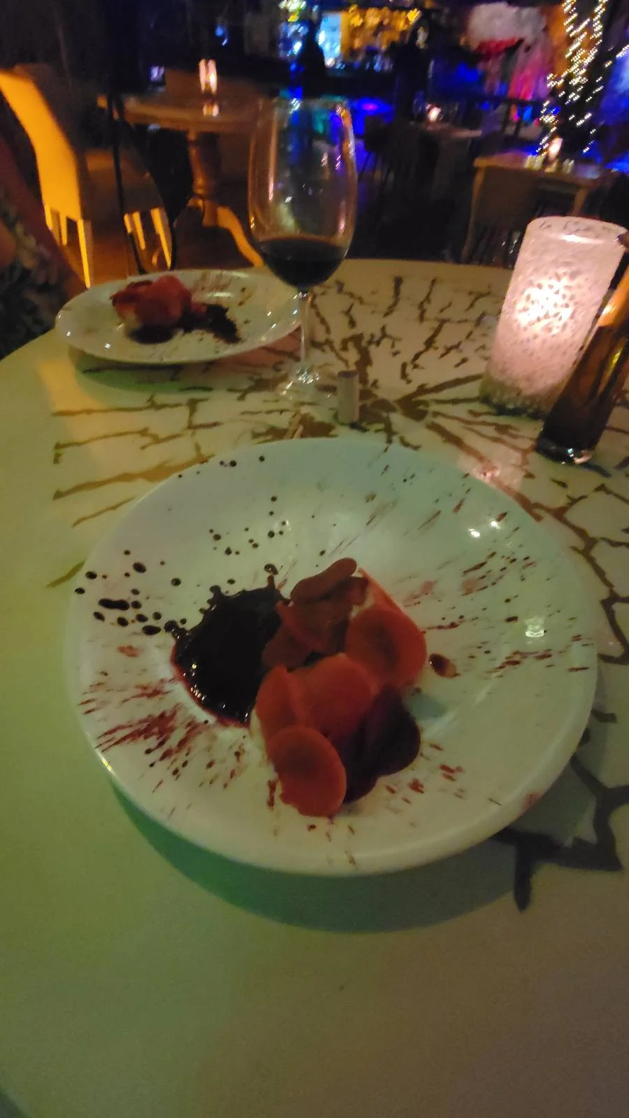Plates with a dessert featuring red drizzles and garnishes on a round table with a lit candle. A glass of red wine is nearby, and there is dim, ambient lighting in the background.