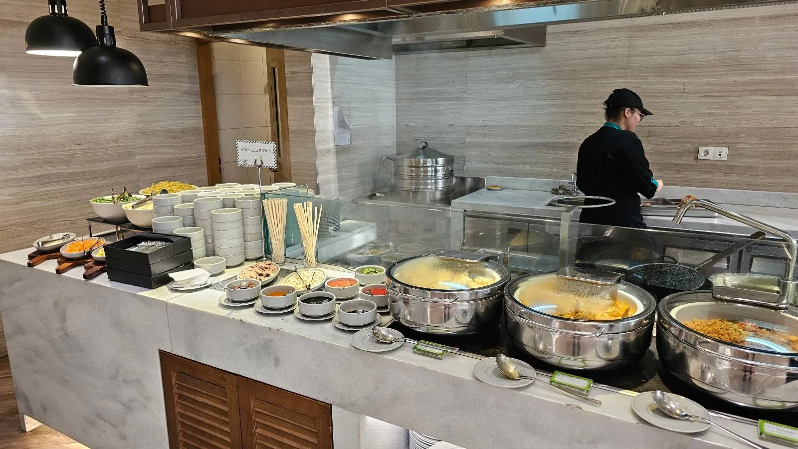 A buffet counter with various dishes in metal chafing dishes, plates, bowls, and utensils neatly arranged. A chef in a black uniform is preparing food behind the counter in a modern kitchen setting.