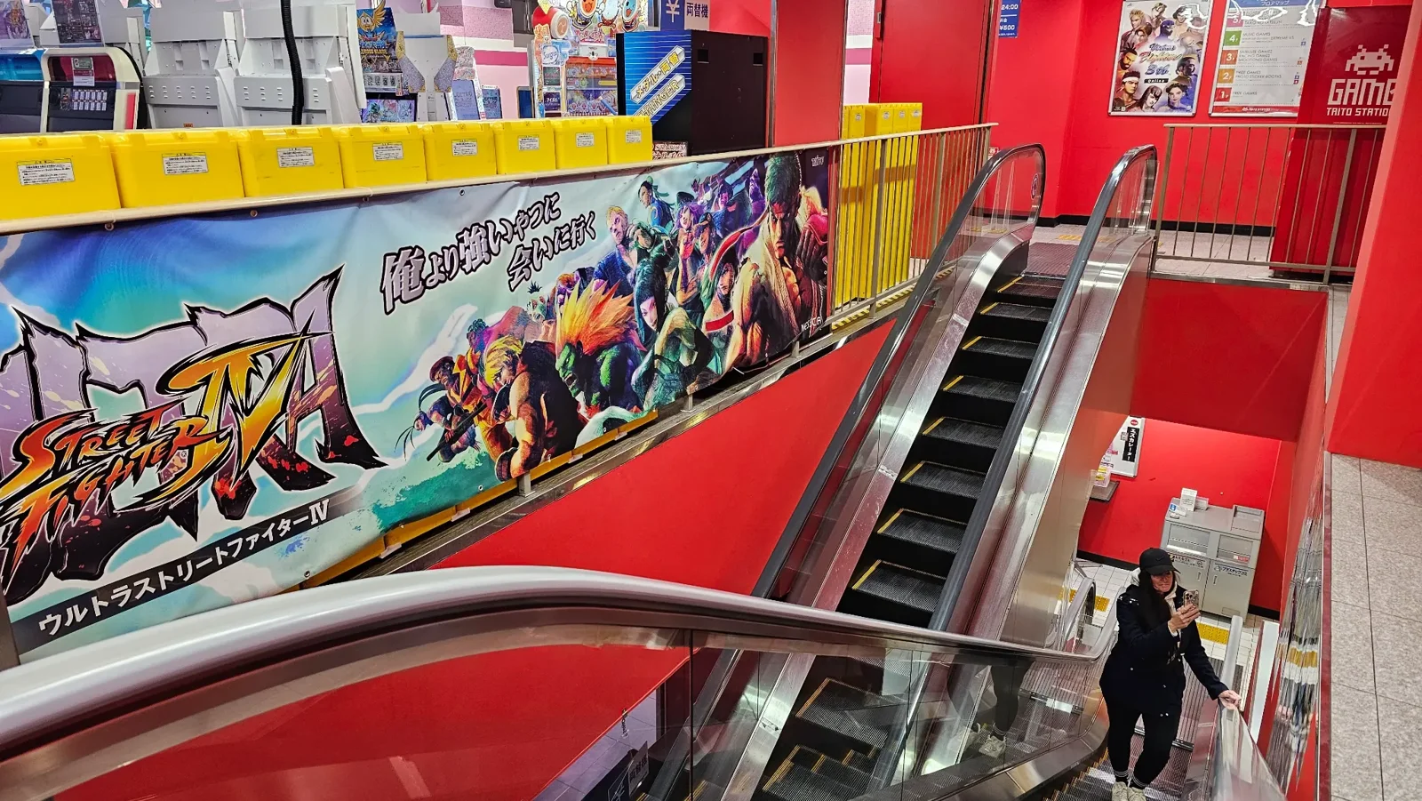 A woman uses an escalator in a brightly lit store with red walls, yellow boxes, and a large colorful "Street Fighter" banner featuring game characters.