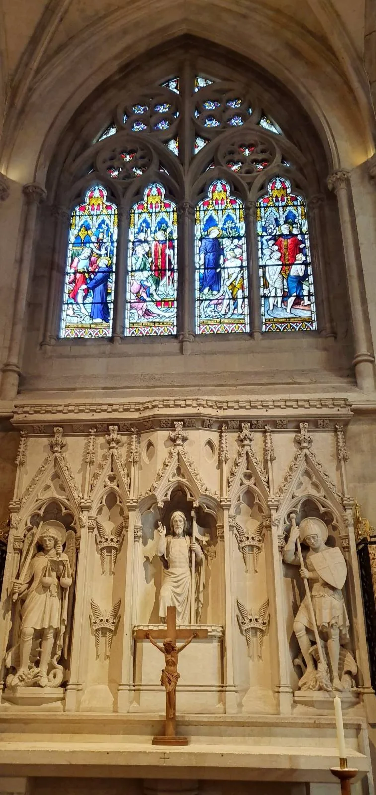 Interior view of a cathedral featuring a high, arched ceiling and a large, intricate rose window above a pipe organ. Rows of wooden pews line the aisle leading to the altar, and ornate chandeliers hang from the ceiling.