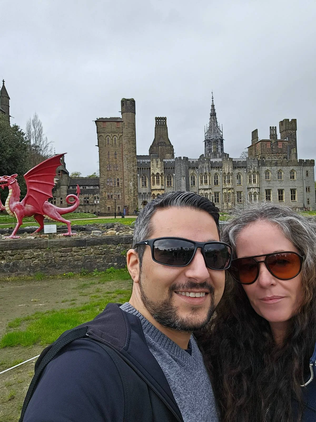 A couple takes a selfie with a red dragon sculpture and a historic castle in the background. both are wearing sunglasses.