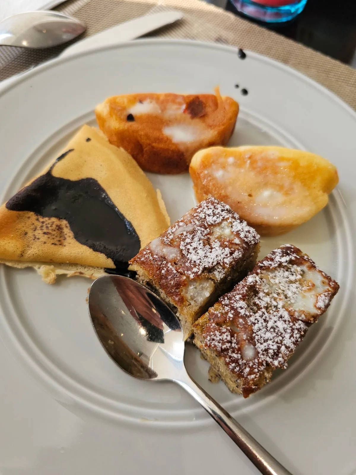 A plate with various desserts: a slice of cheesecake with chocolate drizzle, two pieces of glazed cake, and two fruit-topped tartlets. A spoon rests on the plate.