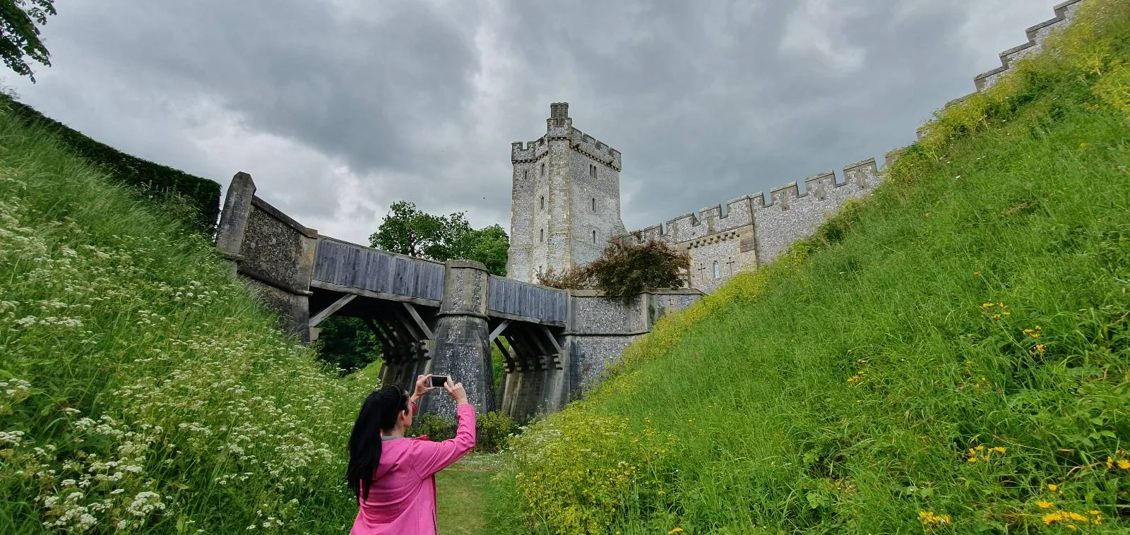 A person in a pink jacket takes a photo of a historic stone castle with a tower and arched wall, set against a cloudy sky. The foreground has sloping grassy hills with wildflowers.