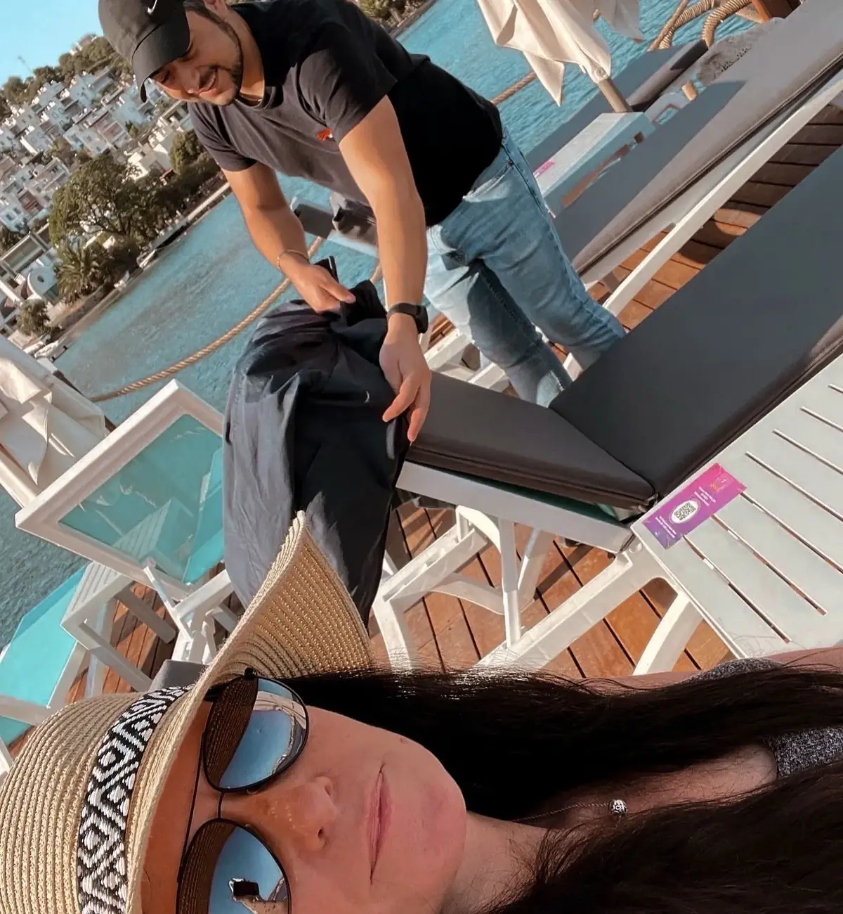 A woman wearing a sun hat and sunglasses takes a selfie on a deck. A man in a cap adjusts a chair in the background near water.