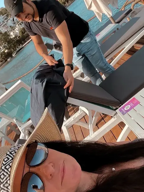 A woman wearing a sun hat and sunglasses takes a selfie on a deck. A man in a cap adjusts a chair in the background near water.