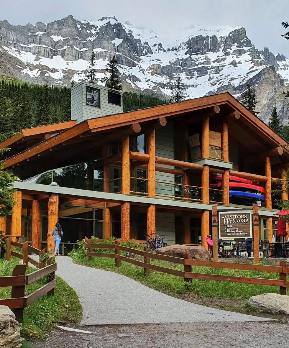 Lodge with wooden beams, backed by mountains. Pathway leads to sign reading "Visitors Welcome." Kayaks stacked nearby, bikes parked.