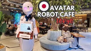 Robot with screen in a cafe setting. Sign reads "Avatar Robot Cafe Tokyo." People sit at wooden tables, greenery decorates the space.