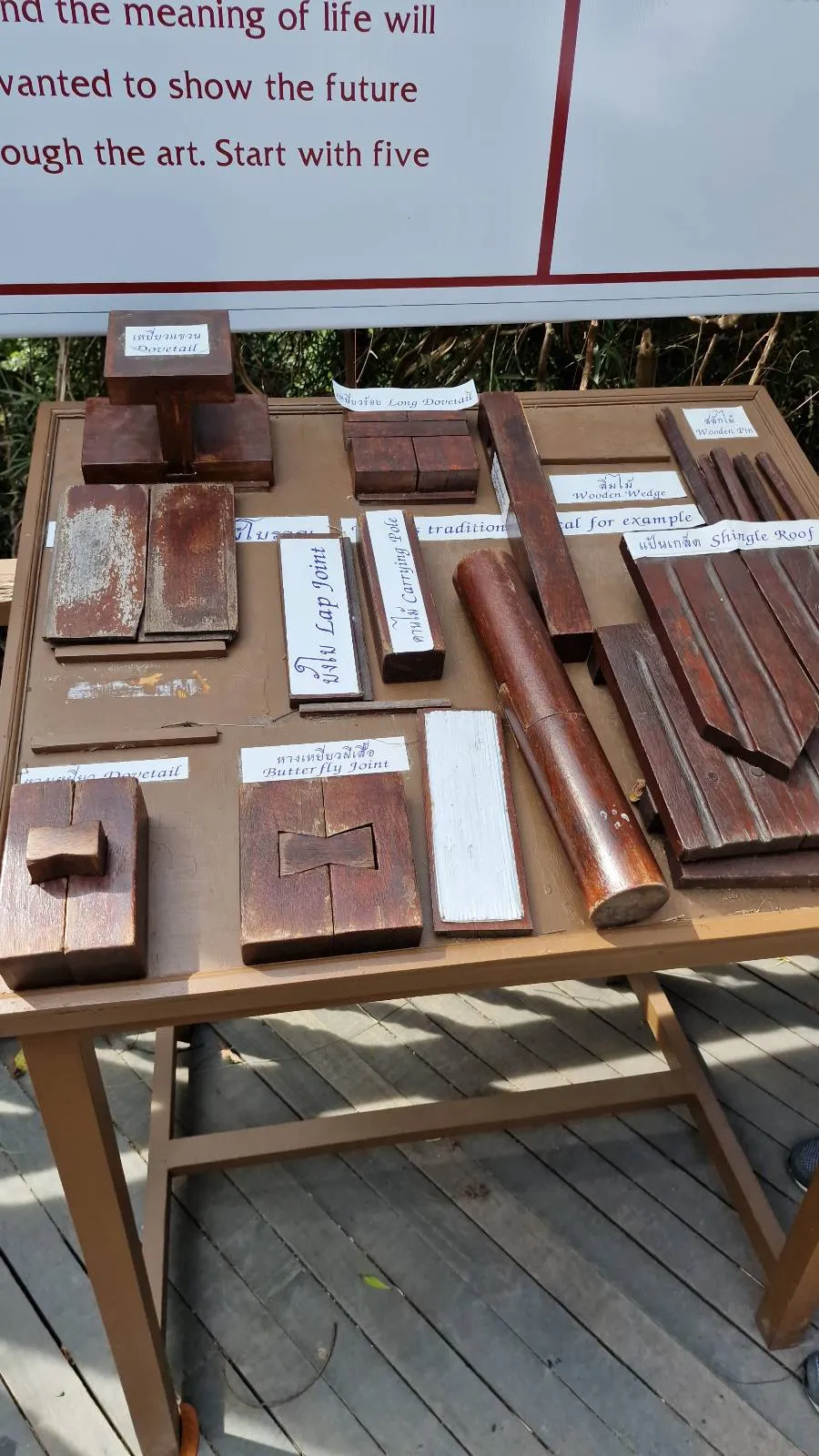 A wooden display table with various rectangular wooden objects arranged on it, each labeled with text. The table is outdoors on a wooden deck, with a banner partially visible in the background.