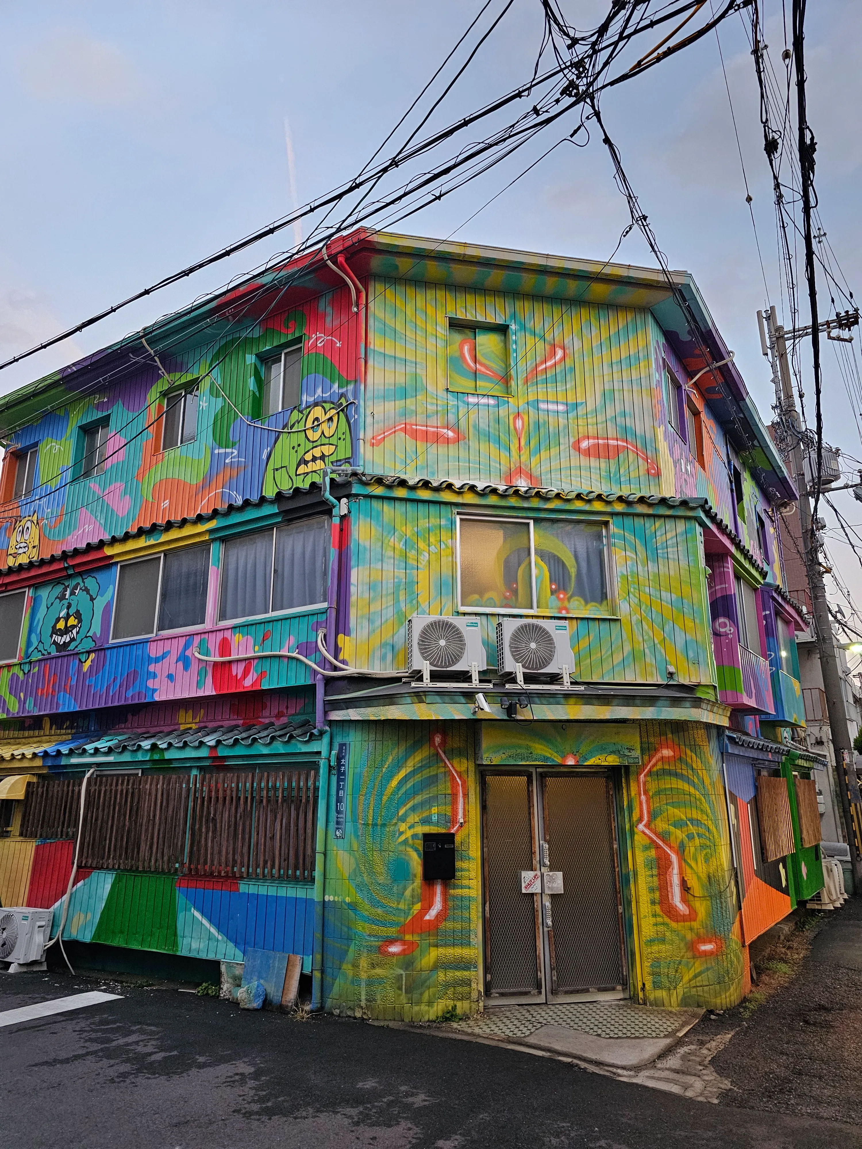 A three-story building painted with colorful, abstract murals, including various geometric shapes and vibrant patterns, stands at a street corner with visible air conditioning units and overhead power lines.
