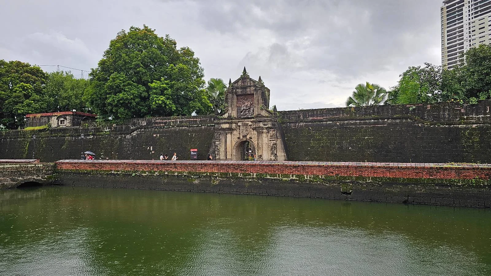 A historic stone fortification with an arched entrance, situated next to a moat. Lush green trees are visible above the structure, and a tall modern building rises in the background under a cloudy sky.