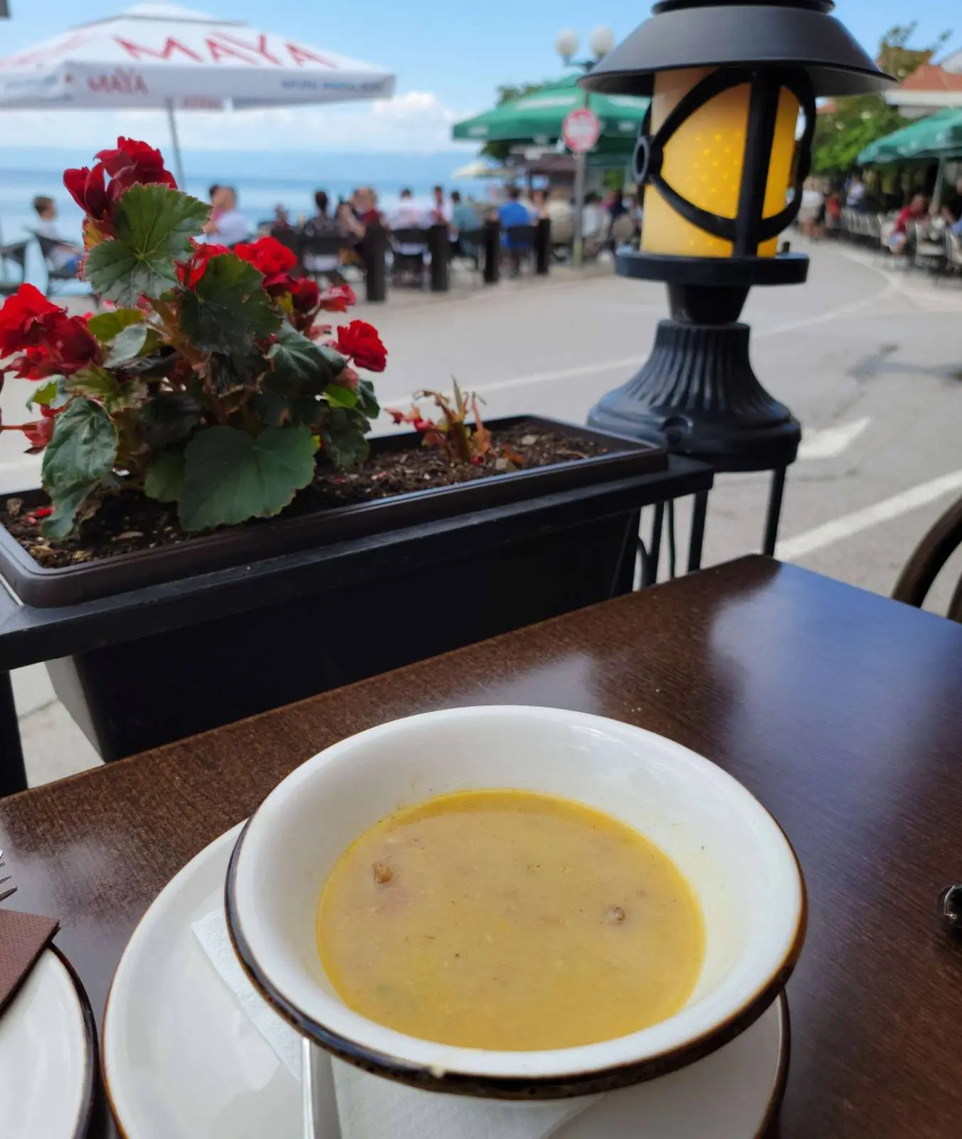 A bowl of soup sits on a dark wooden table at an outdoor café. Red flowers are in a planter nearby, and a decorative lamp is on the railing. In the background, there are tables, chairs, and an umbrella with people enjoying a waterfront view.