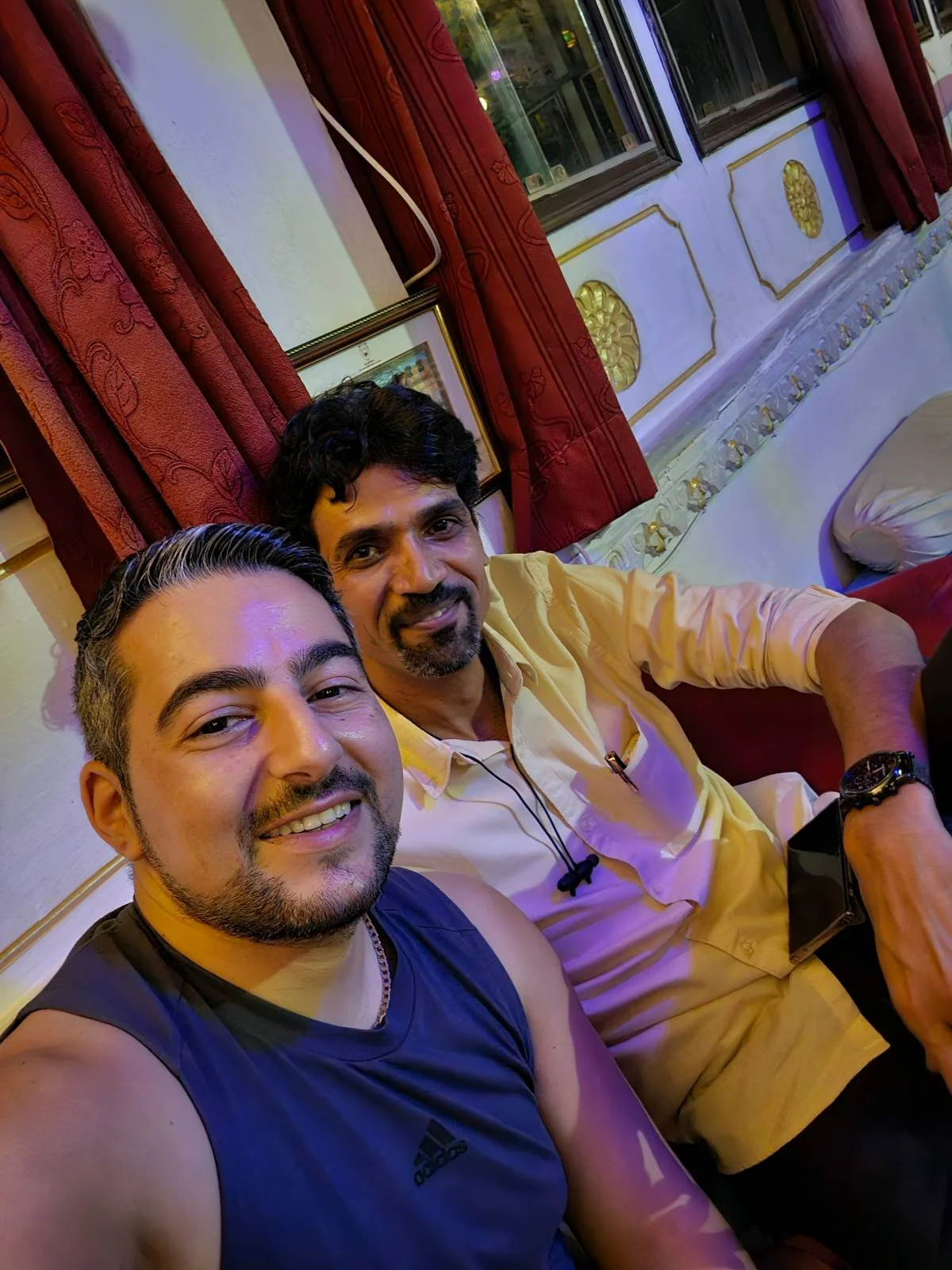 Two men sit indoors on a couch; one in a dark sleeveless shirt smiles at the camera, while the other in a light-colored shirt sits next to him. Red curtains and ornate decor are visible in the background.