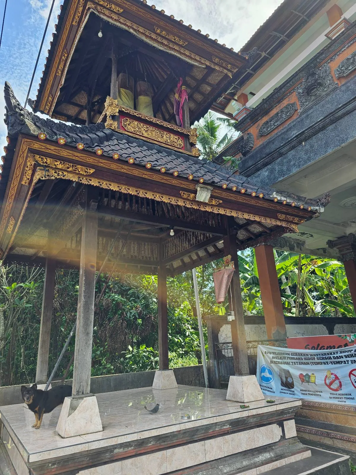 A small, elevated pavilion with a traditional Balinese roof. Sunlight filters through, creating rays on the structure. Lush greenery and part of another building are visible in the background. A barge with a poster is near the pavilion.