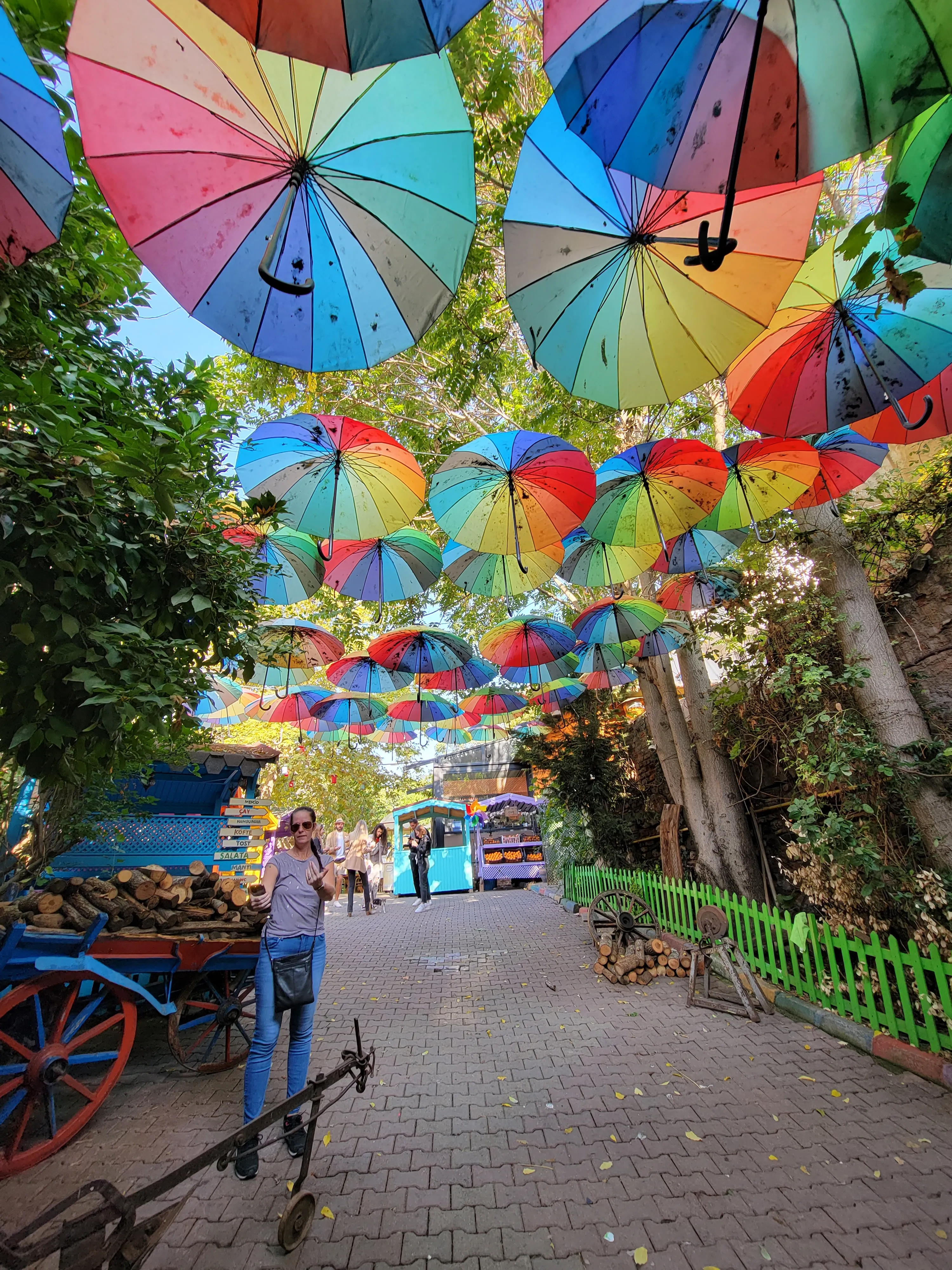A walkway is decorated with colorful umbrellas hanging overhead. Trees line the path, and a person is walking along the path near a blue and orange cart. The atmosphere is vibrant and festive.