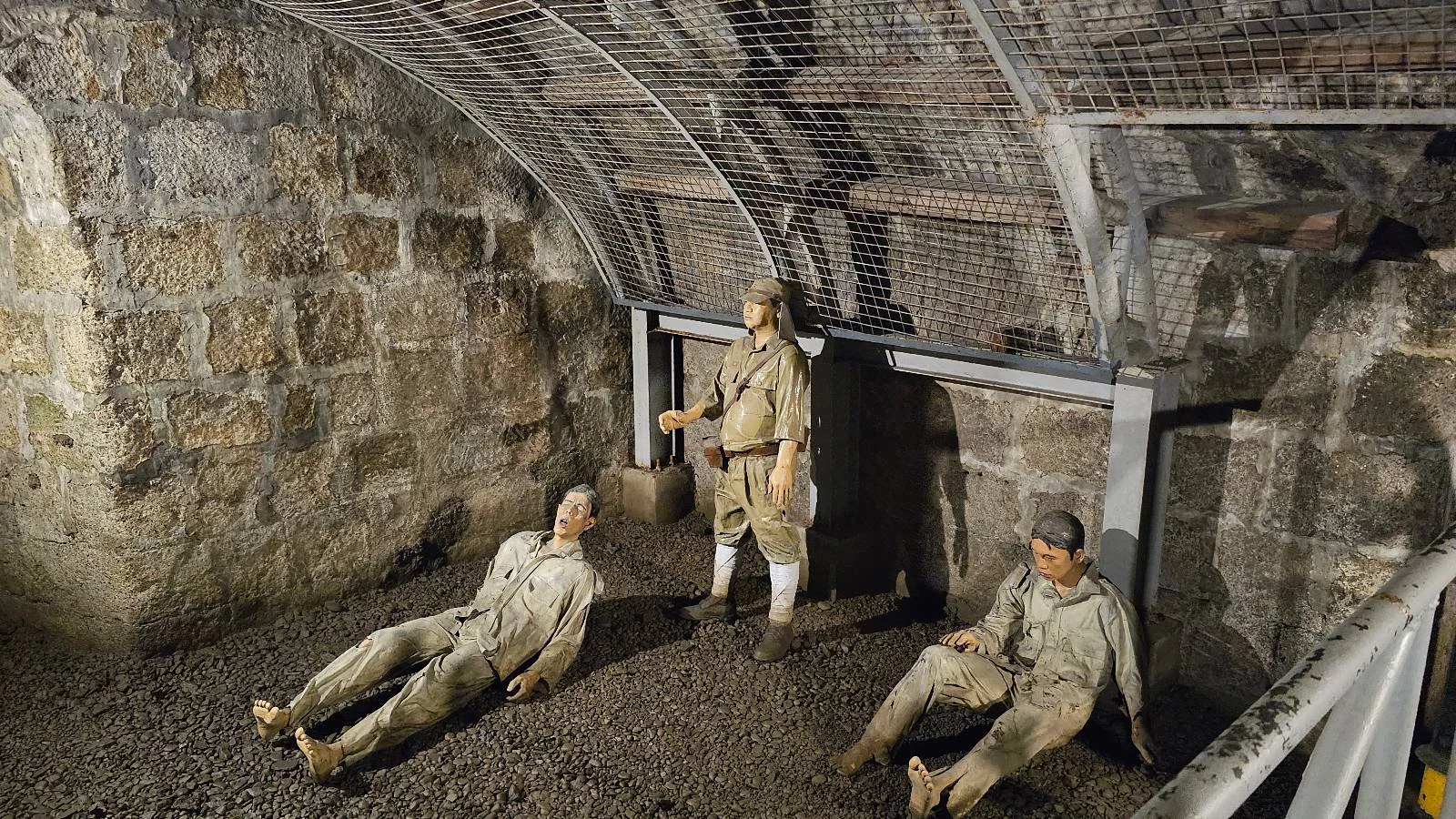 A historical display featuring three mannequin soldiers in military uniforms sitting and standing on gravel in a dimly lit underground stone bunker with arched ceilings. Two are seated against the walls, and one is standing.