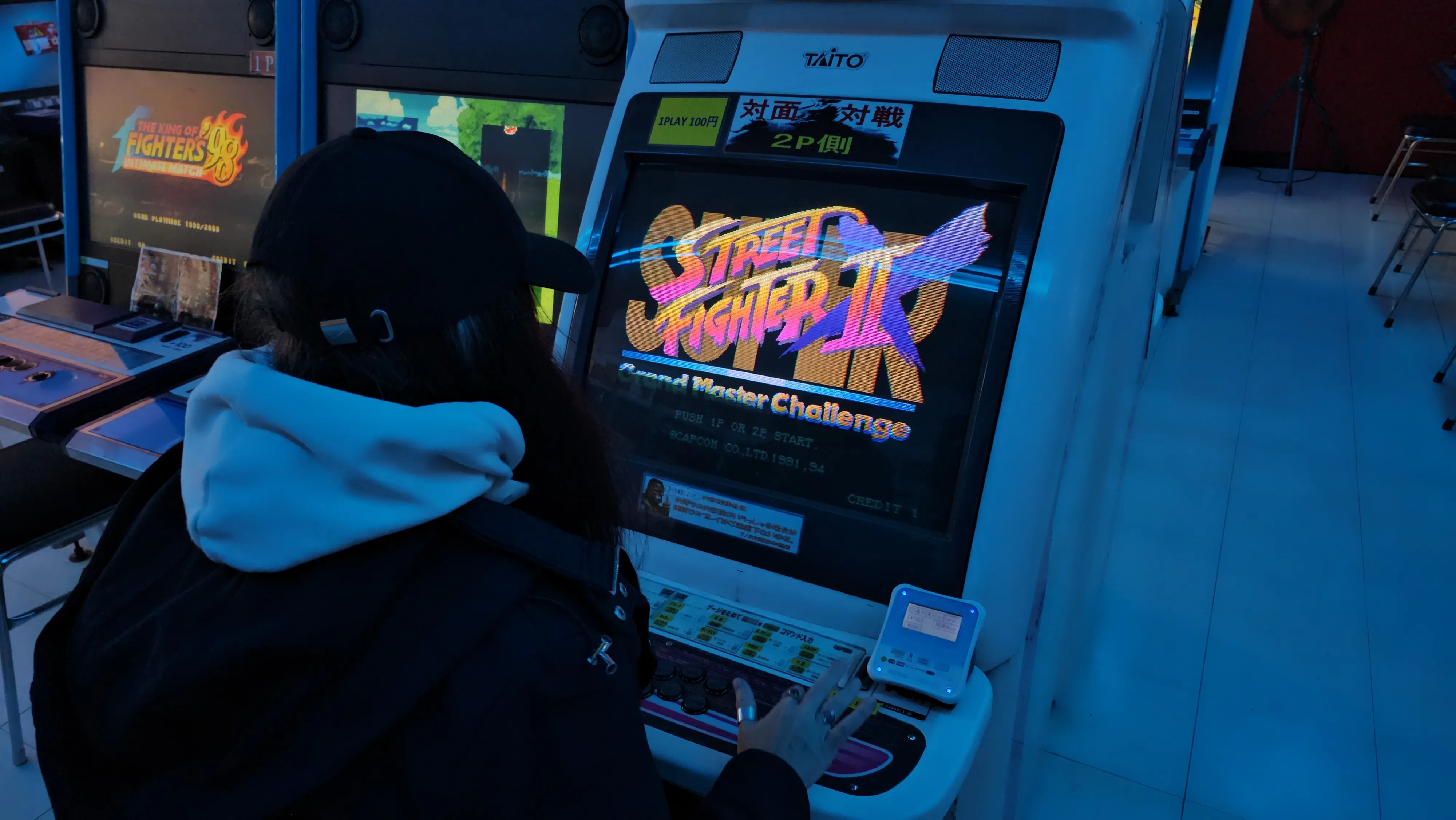 A person in a hoodie and cap plays Street Fighter II on an arcade machine in a dimly lit gaming area.