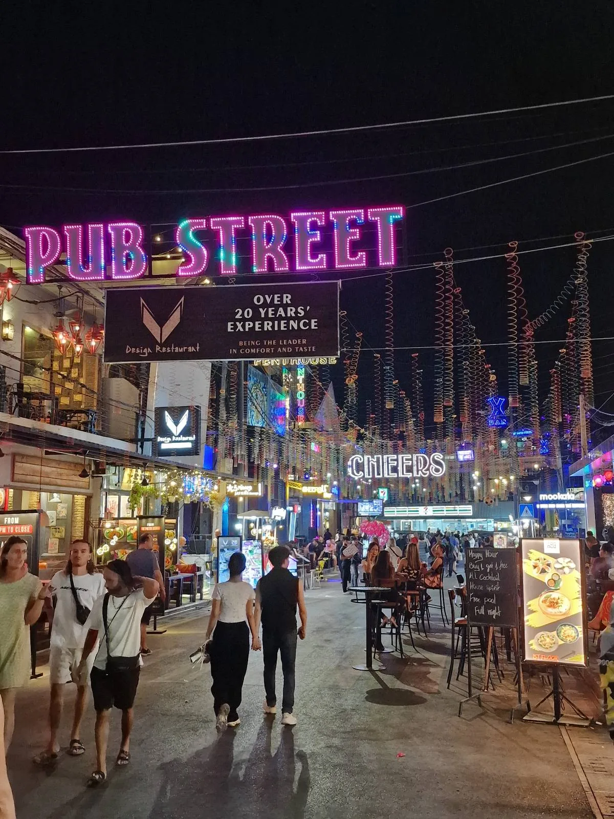 A vibrant street scene at night with people walking under a glowing "PUB STREET" sign, surrounded by brightly lit bars, restaurants, and colorful lights.