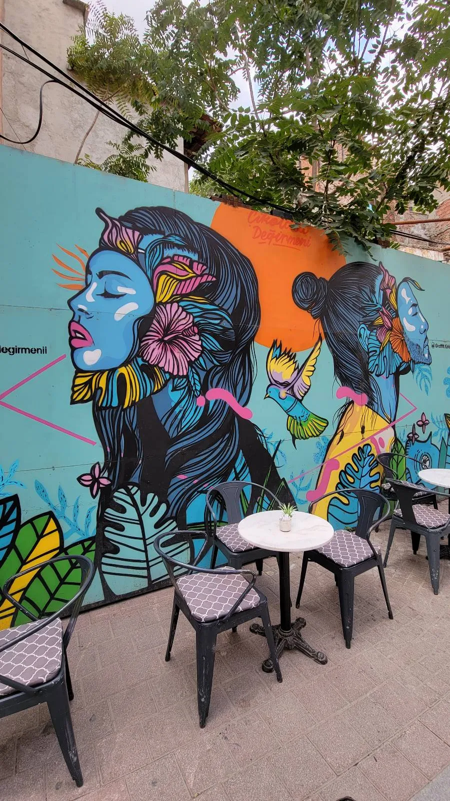 Colorful mural on an outdoor wall depicts two stylized female faces with vibrant blue and pink accents. Tables and chairs are arranged in front, and trees are visible above the wall.