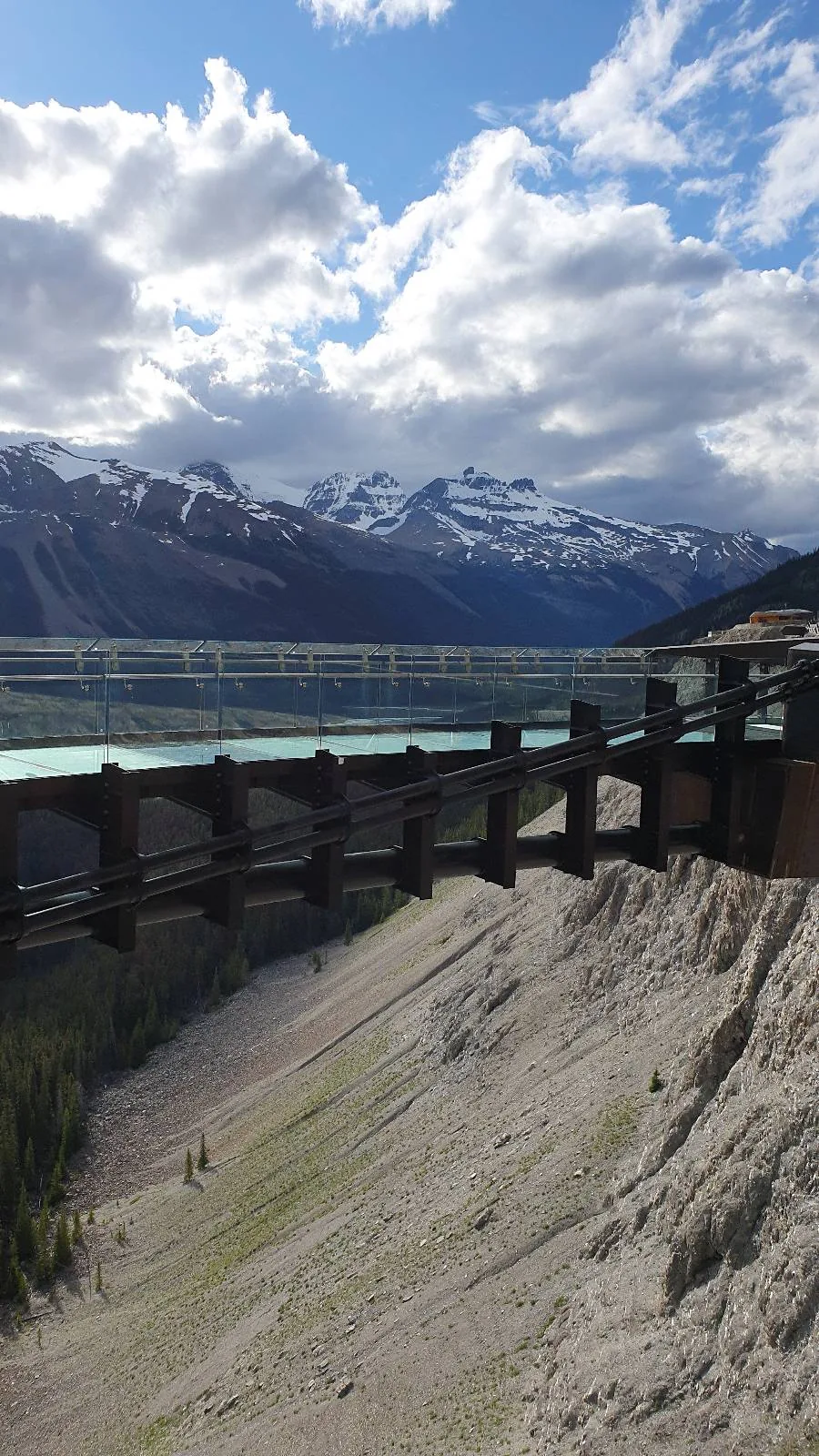 Wooden railing overlooks a turquoise river and pine trees, with snow-capped mountains and a partly cloudy sky in the background. A building is partially visible on the right side.