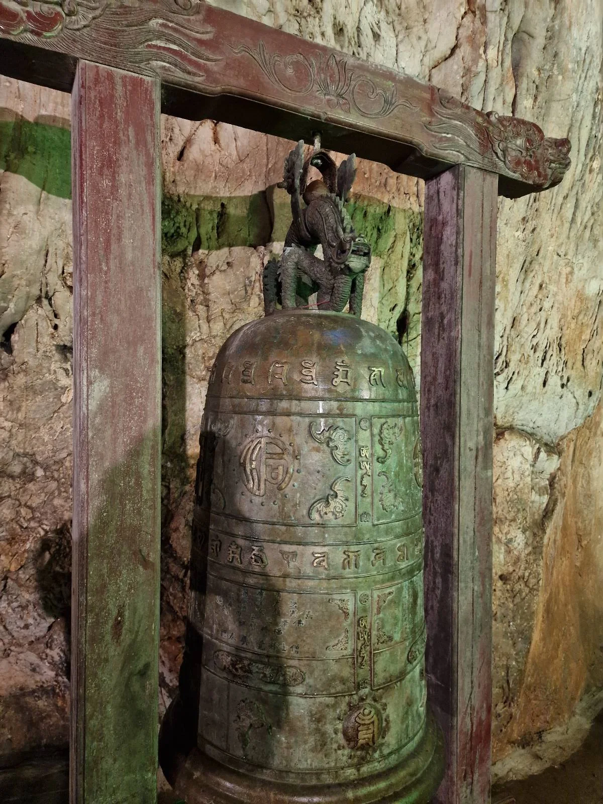 Ancient bell with intricate carvings hanging under a wooden frame inside a cave.