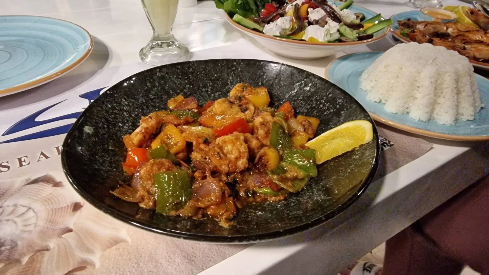 A black bowl containing a dish made of mixed vegetables and meat, garnished with a lemon wedge, is placed on a table next to a small mound of rice and a salad.