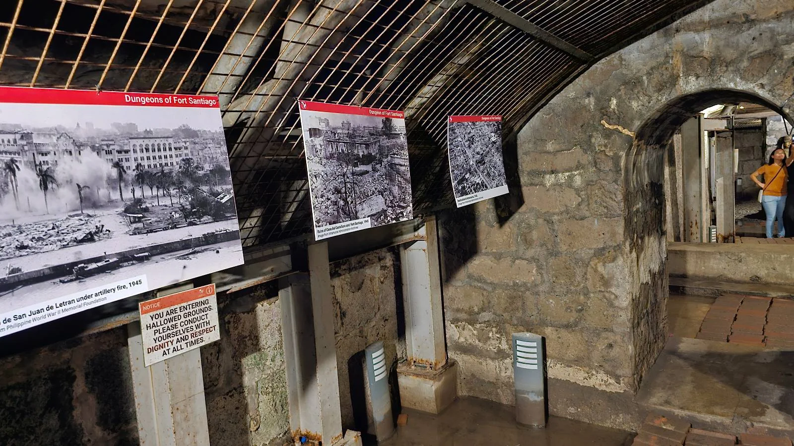 An underground tunnel with stone walls displays historical photographs and informational signs. A person is visible in the background, walking along the path beneath a metal grate ceiling.
