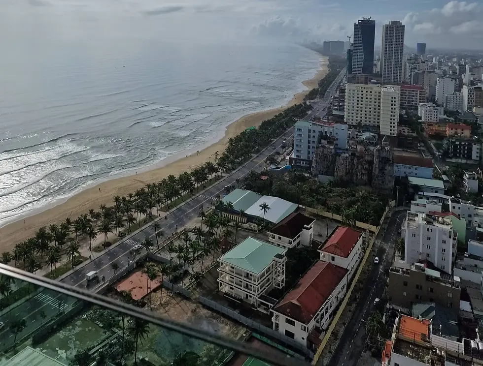Aerial view of a coastal cityscape with high-rise buildings, a road lined with palm trees, and a sandy beach adjacent to the calm ocean.