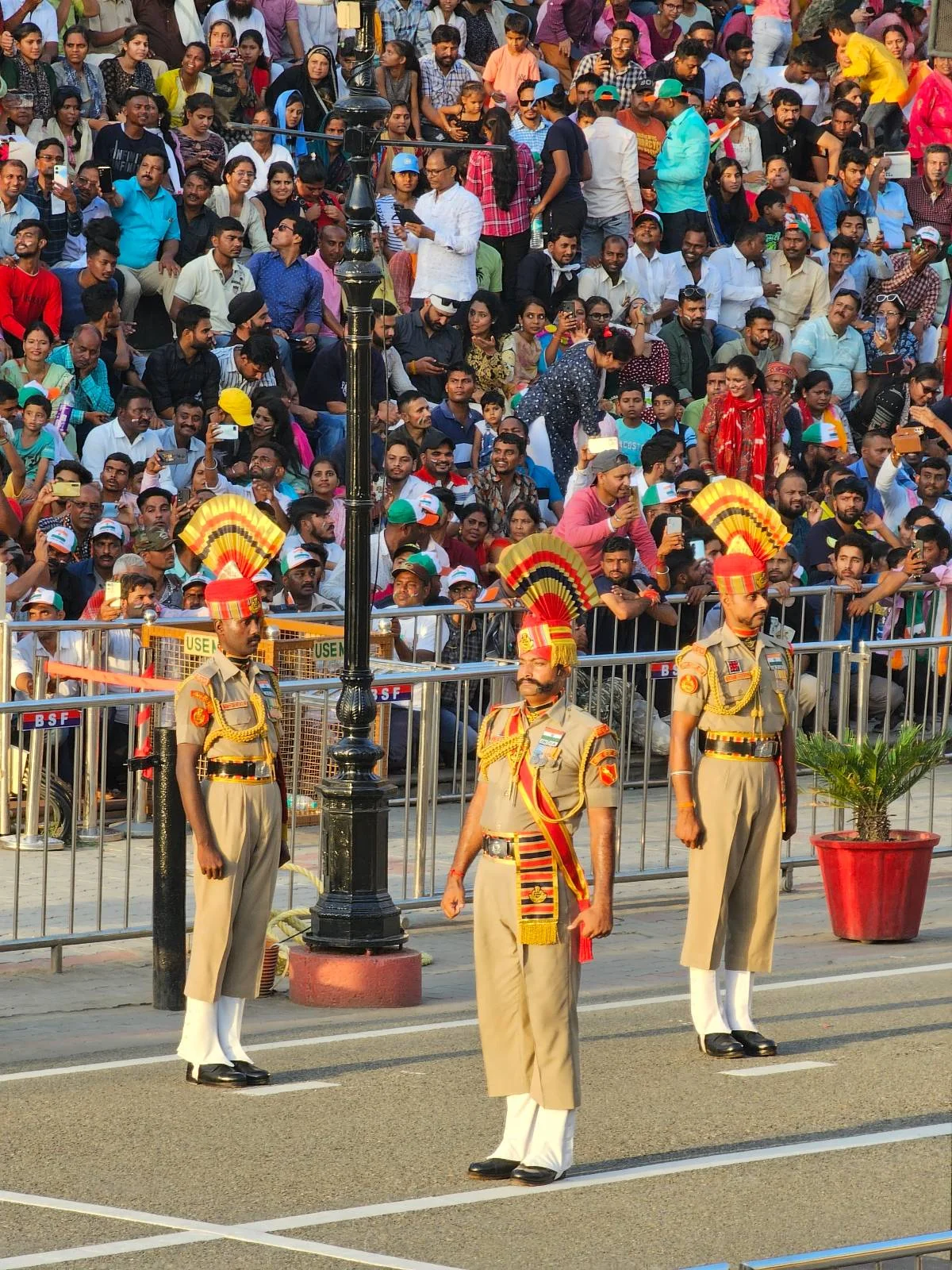 Three uniformed soldiers in ceremonial attire stand at attention in front of a large crowd, with colorful hats and sashes, participating in a public event or parade with a street and fence in the background.