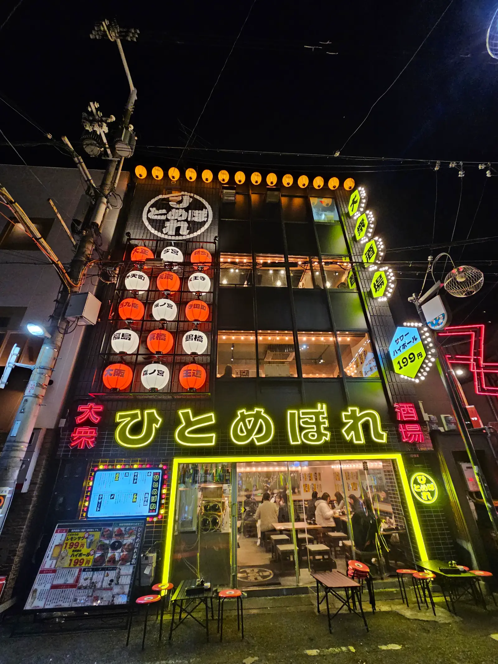 A vibrant, multi-story building in Japan is lit with colorful neon signs and lanterns at night. The ground-floor windows reveal diners inside, and the glowing signage features Japanese characters in yellow, red, and green.