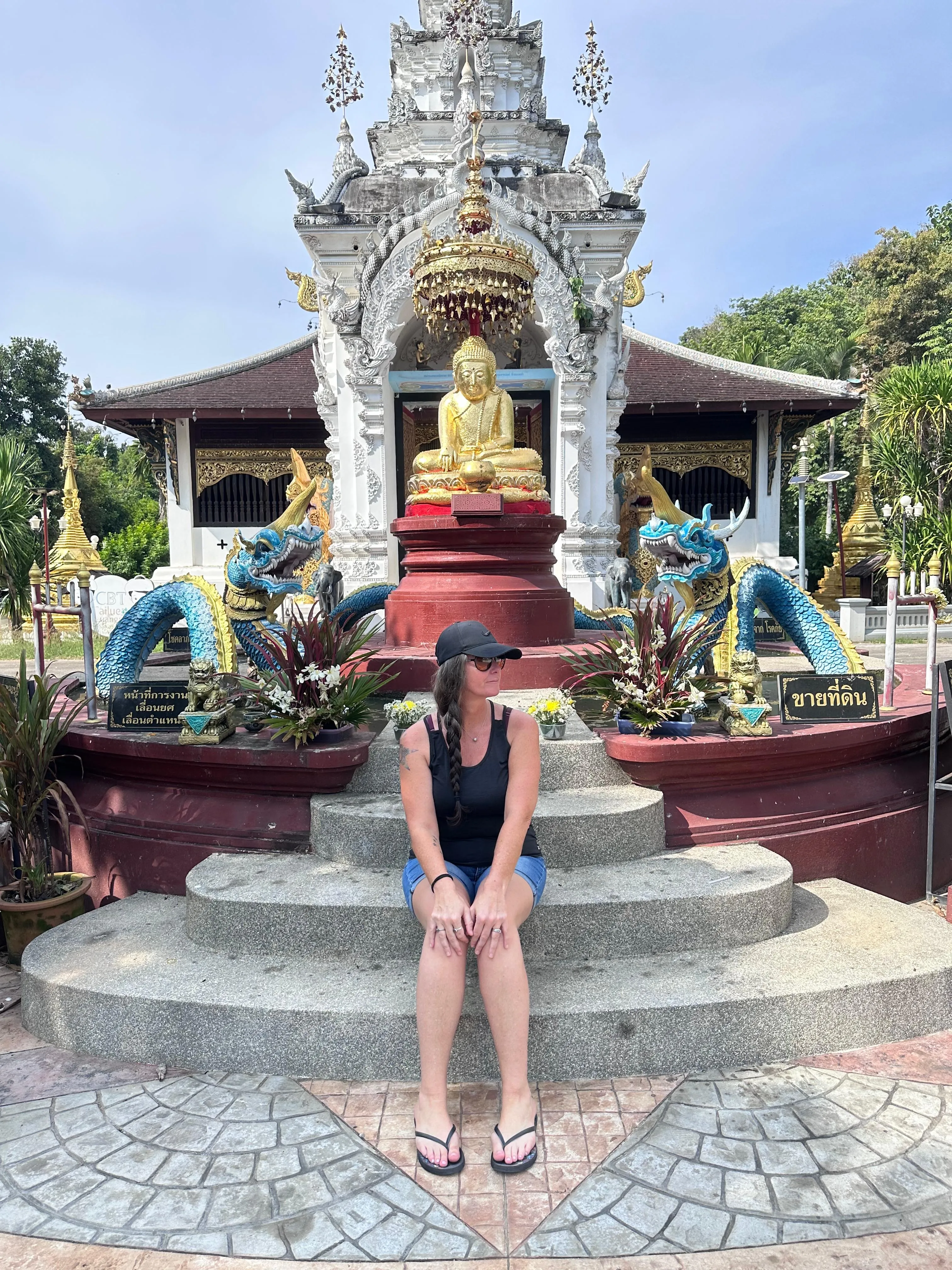 A woman in a black tank top and shorts sits on stone steps in front of a golden Buddha statue with ornate decorations and dragon sculptures at an outdoor temple.