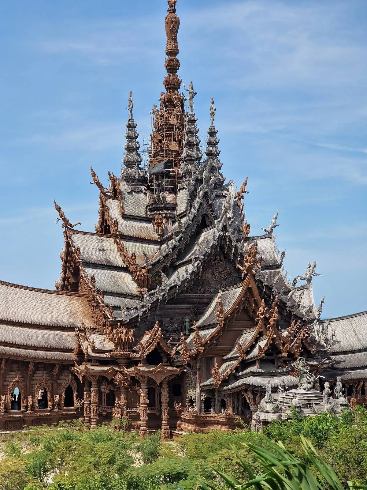 A large, intricately carved wooden temple with multiple tiers and spires, set against a blue sky. The structure features detailed carvings and sculptures, showcasing traditional architectural style. Lush greenery surrounds the temple.