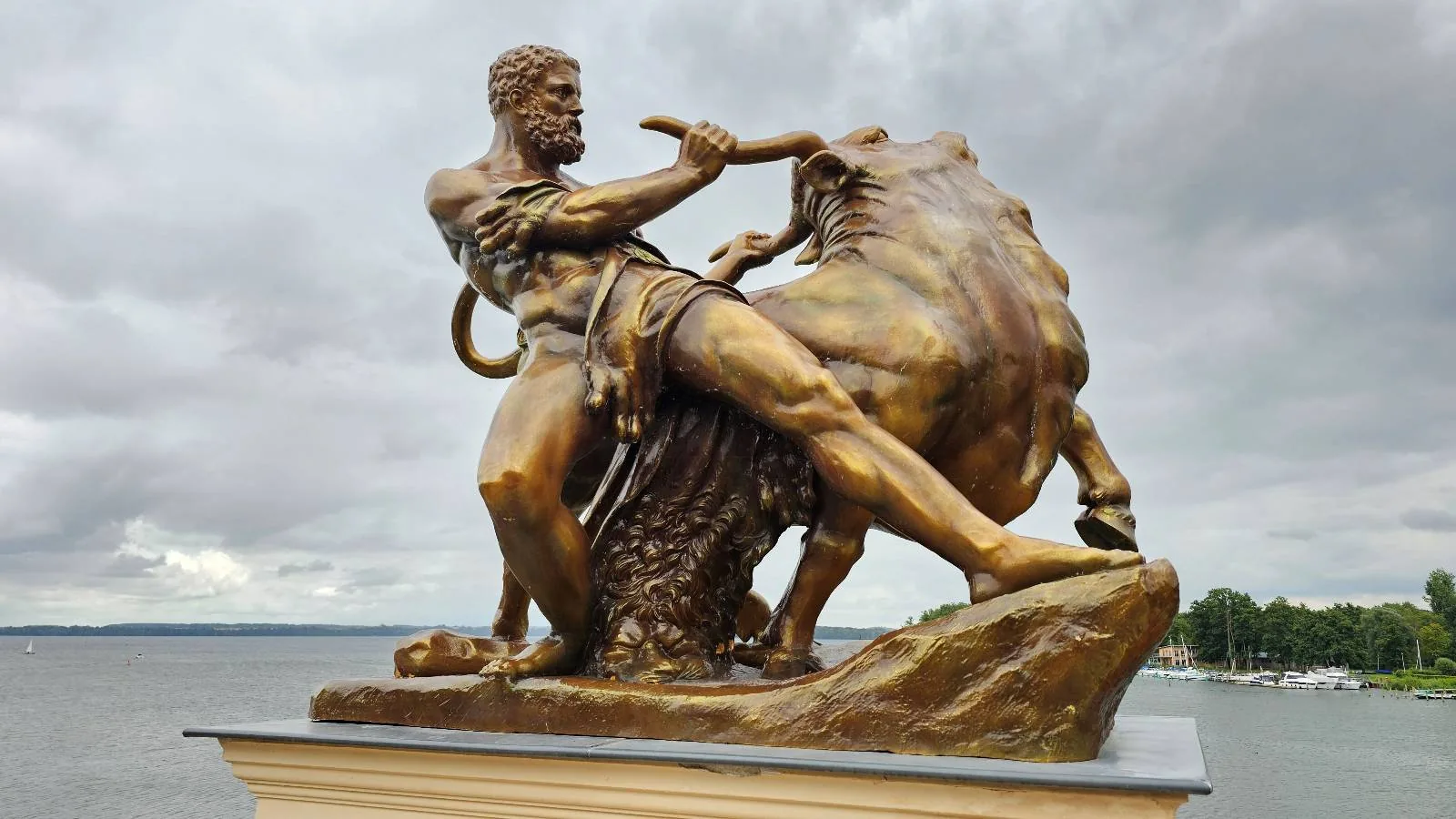 A bronze statue of a muscular man wrestling a lion stands on a pedestal by the water, with a cloudy sky in the background.