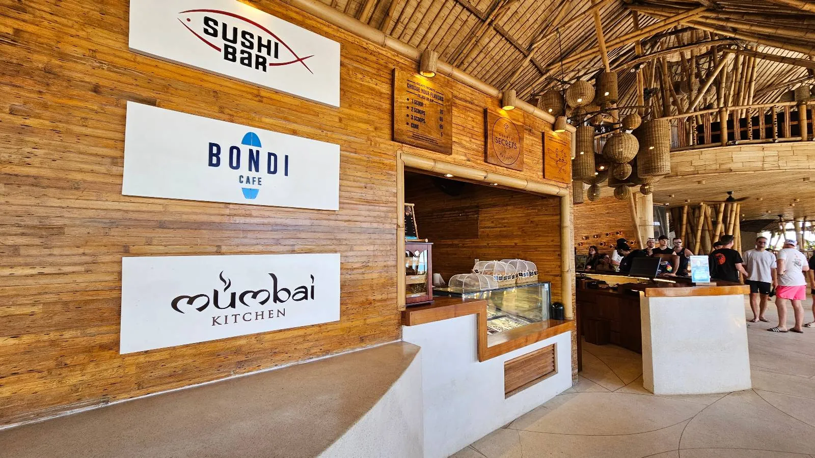 Interior of a restaurant with wooden decor, featuring three food counters labeled "Sushi Bar," "Bondi Grill," and "Mumbai Kitchen." People are seen in the background. The setting is open and airy with natural lighting.