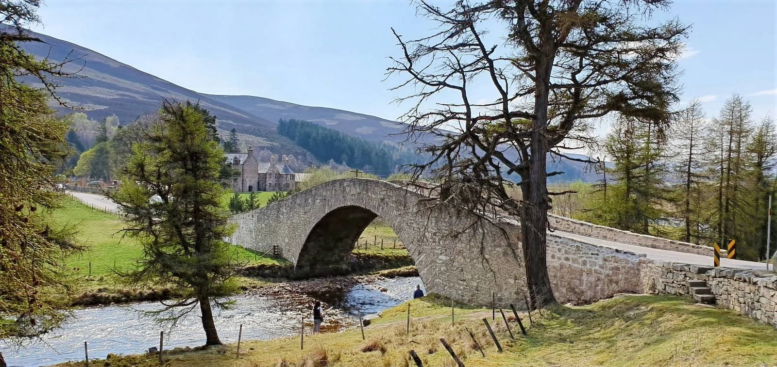 Stone bridge over a stream, surrounded by trees and mountains. A person stands by the water, and stone buildings are in the background.