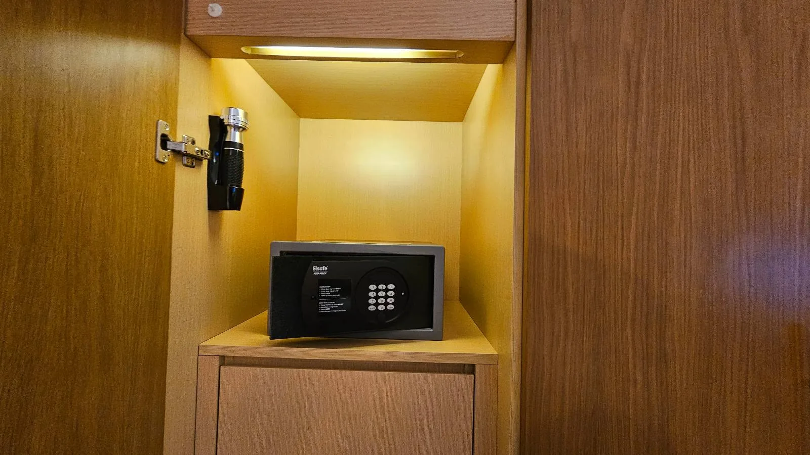 A small black electronic safe with a keypad and display is placed on a wooden shelf inside a built-in cabinet, illuminated by a light above.