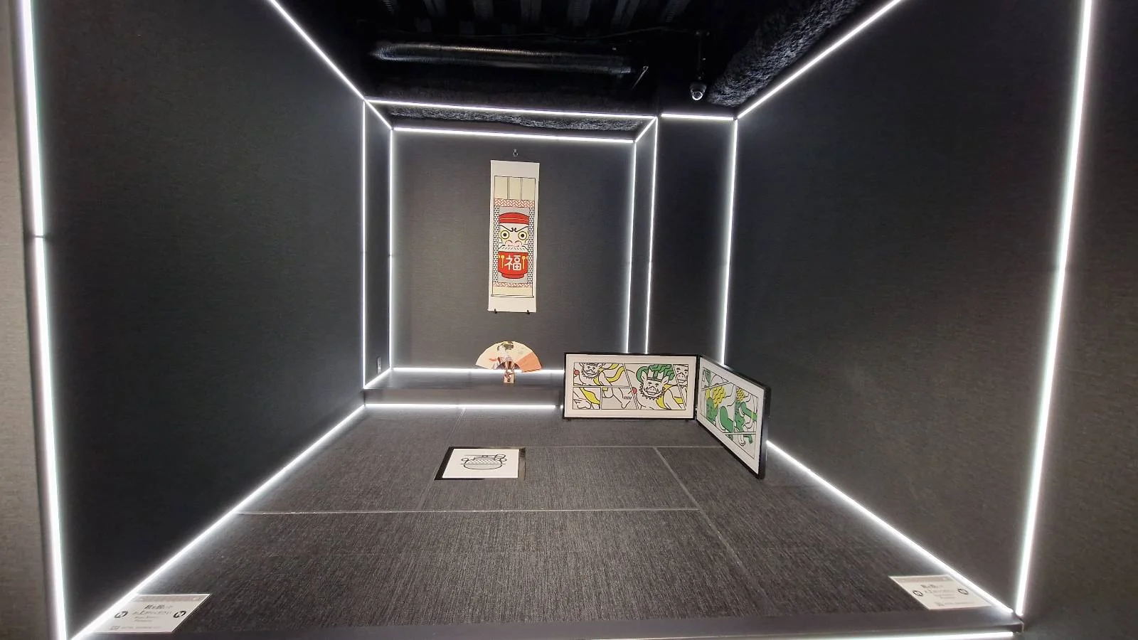 A black room with white LED strip lighting outlines. Inside, there's a poster with abstract art on the far wall and two artworks with geometric patterns on the floor. A small, colorful object lies in the corner.