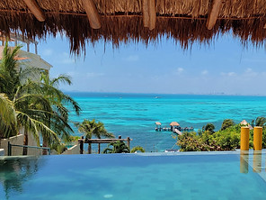 A picturesque view of a tropical paradise featuring an infinity pool overlooking the vibrant blue ocean. Palm trees line the shore, and a thatched roof hangs above, framing the serene, sunny scene reminiscent of Mexico's stunning beaches. Small piers extend into the water.