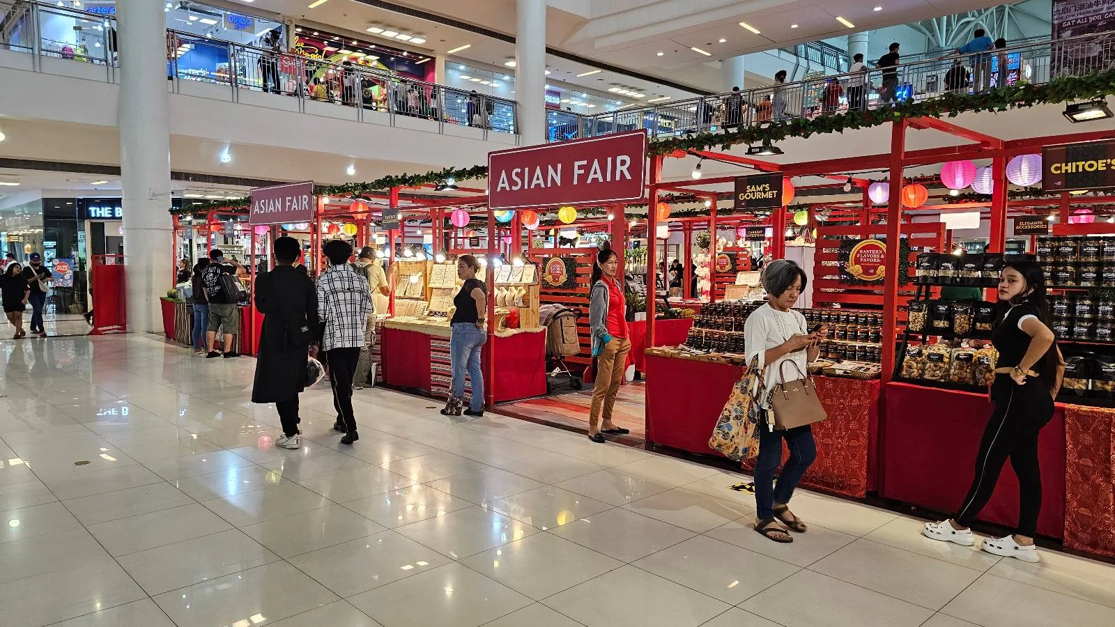 A busy indoor market scene featuring a series of red stalls labeled "Asian Fair." Shoppers browse and interact with vendors. The area is brightly lit, and people are dressed in casual attire, creating a vibrant and lively atmosphere.