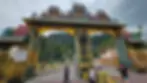 Colorful ornate temple gate with sculptures; people walking below. Lush hills in the background. Sign reads: "Gate No. 1, Opening Time Daily."