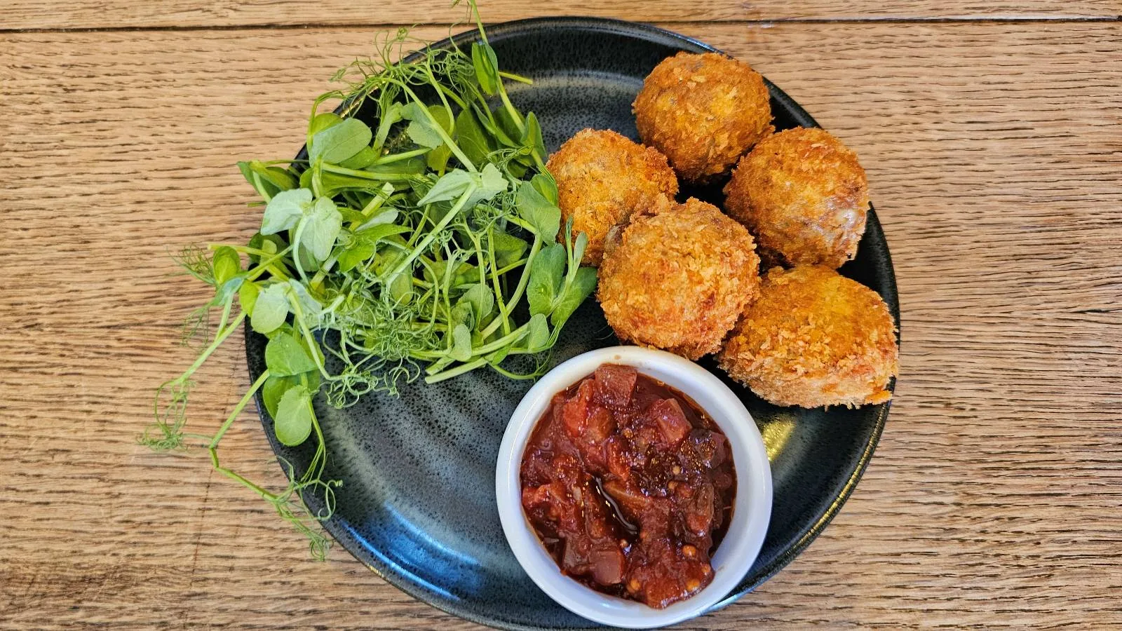 A plate with five golden-brown breaded balls, a serving of fresh green salad, and a small bowl of red sauce, all arranged neatly on a dark plate on a wooden table.